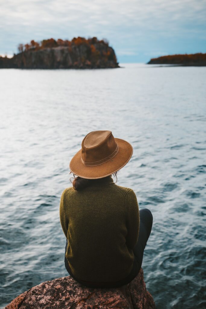 A visitor sitting on a rock at the water's edge, looking out to a wooded island